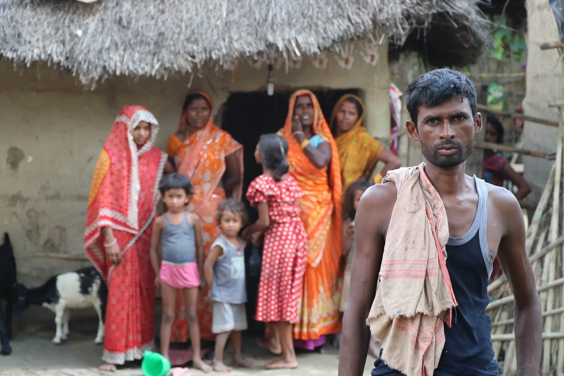 Women and girls outside a thatched home in a village, a man standing in the foreground.